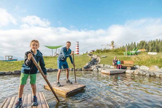 Zwei Kinder stehen auf einer Holzplattform im Wasser und haben Spaß. Im Hintergrund sind grüne Wiesen und ein Leuchtturm zu sehen.