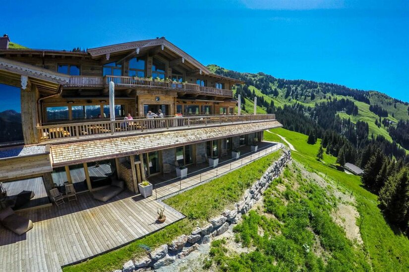 BichlAlm im Sommer Große Berghütte aus Holz mit Sonnenterasse vor bewaldeten Berghängen unter blauem Himmel.