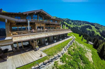 BichlAlm im Sommer Große Berghütte aus Holz mit Sonnenterasse vor bewaldeten Berghängen unter blauem Himmel.