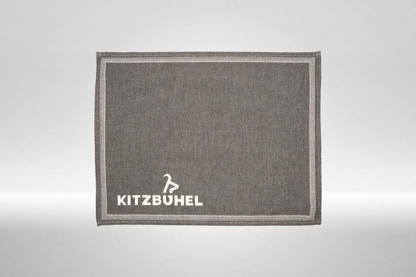 Ein graues Platzset mit einem weißen Rand und dem Schriftzug 'KITZBÜHEL' in der unteren rechten Ecke.