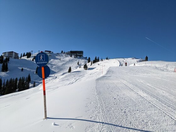 Winterwanderweg im Skigebiet Präparierter Winterwanderweg auf einer verschneiten Anhöhe mit blauem Hinweisschild unter klarem Himmel.