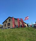 Steinerne Berghütte mit Terrasse, Fahnen und Mountainbikes auf einer grünen Wiese unter blauem Himmel.