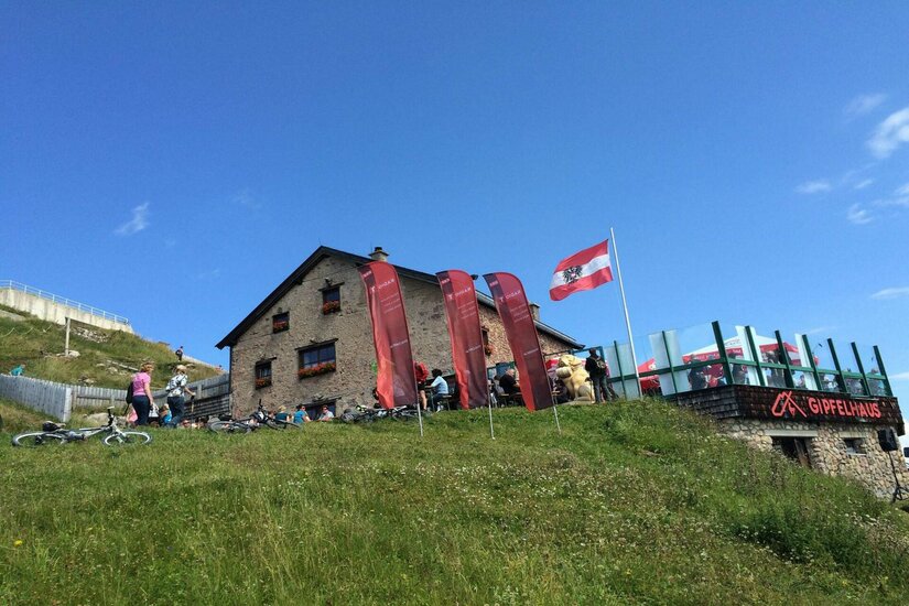 Gipfelhaus im Sommer Steinerne Berghütte mit Terrasse, Fahnen und Mountainbikes auf einer grünen Wiese unter blauem Himmel.