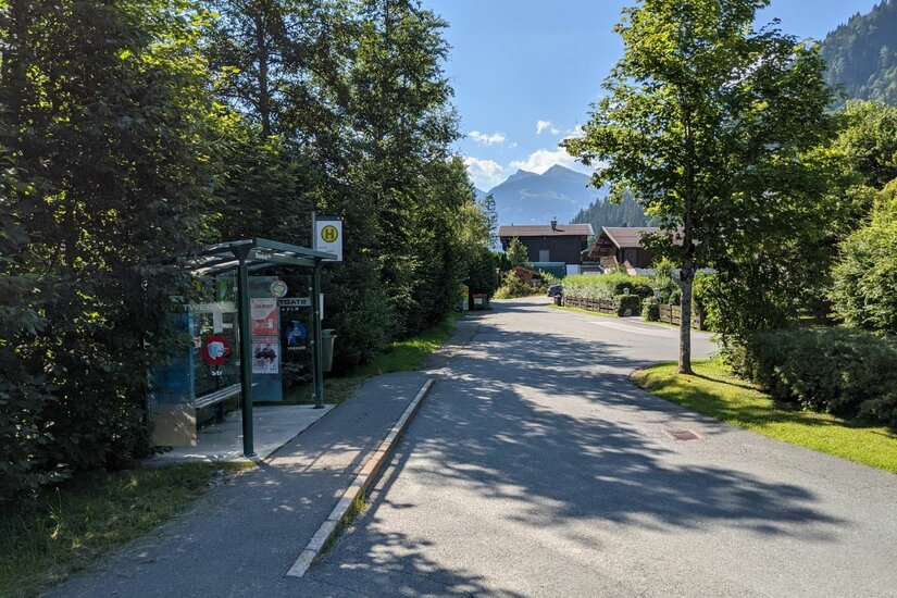 Bushaltestelle mit Wartehäuschen an einer asphaltierten Straße in Staudach vor Bergkulisse.