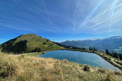 Ein künstlicher Speichersee auf einem Bergrücken vor grasbewachsenen Gipfeln und blauem Himmel.