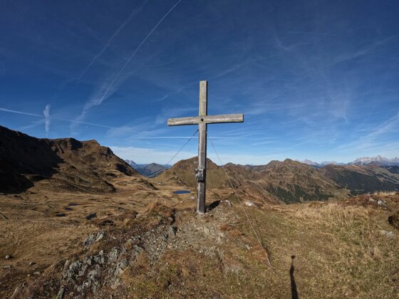 Gipfelkreuz am Mittagskogel Ein hölzernes Gipfelkreuz auf einem grasbewachsenen Bergrücken vor einem Panorama aus braunen Alpengipfeln.