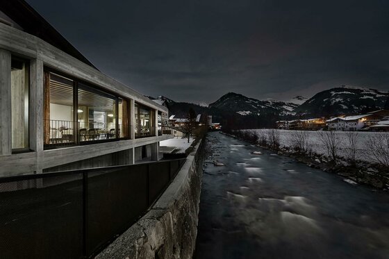 Auwirt im Winter Modernes Gebäude mit großen Glasfronten bei Nacht neben einem Fluss in winterlicher Berglandschaft.