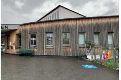 Behindertenparkplatz - Im Grieß 13 Ein modernes Gebäude mit Holzfassade und großen Fenstern. Vor dem Eingang sind Sitzgelegenheiten und ein Parkplatz für Menschen mit Behinderung.