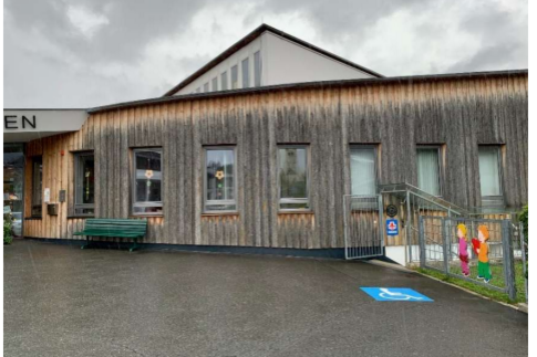 Behindertenparkplatz - Im Grieß 13 Ein modernes Gebäude mit Holzfassade und großen Fenstern. Vor dem Eingang sind Sitzgelegenheiten und ein Parkplatz für Menschen mit Behinderung.