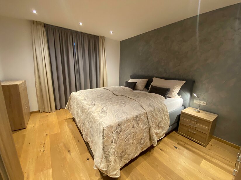 Ein modernes Schlafzimmer mit einem Doppelbett und Holzfußboden. Die Wände sind in neutralen Farben gehalten, und es gibt Vorhänge sowie eine Kommode.
