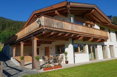 Haus Ansicht Sommer Zweistöckiges alpines Wohnhaus mit Holzfassade, Balkon und Terrasse vor einer Wiese.