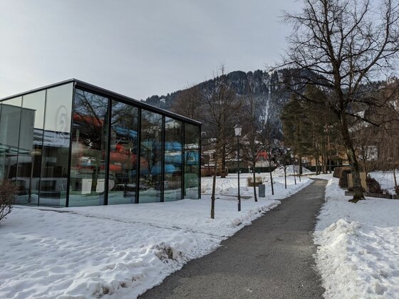 Modernes Glasgebäude neben einem geräumten Fußweg in einer verschneiten Parkanlage vor bewaldetem Berg.