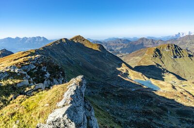 Blick von einem felsigen Berggipfel auf einen Gebirgskamm und einen kleinen Bergsee unter blauem Himmel.