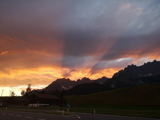 Abendstimmung am Wilden Kaiser Silhouette eines gezackten Gebirgsmassivs vor einem orange-violett gefärbten Abendhimmel mit Wolkenstrahlen.