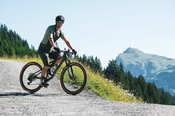 Mountainbiker auf Schotterweg Ein Mountainbiker fährt auf einem Schotterweg vor einer Bergkulisse mit Wald.