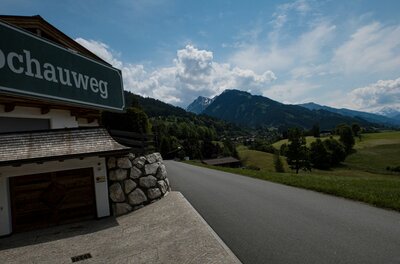 Asphaltierte Straße führt an einem Gebäude mit der Aufschrift Kochauweg vorbei in eine Berglandschaft.