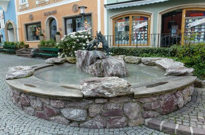 Brunnen mit spielenden Gemsen Ein schöner Brunnen mit einer Statue auf einem Felsen. Umgeben von Steinen und einer gepflasterten Straße.