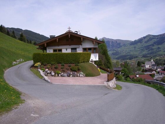 Haus Schuh am Hang Alpines Wohnhaus an einer asphaltierten Kurve mit Blick auf ein Dorf im Tal und bewaldete Berge.