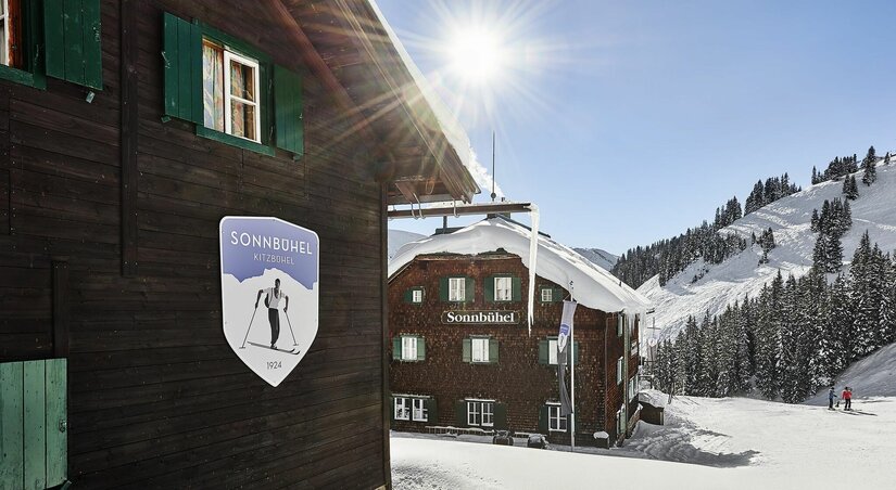Berghütte Sonnbühel in Kitzbühel Holzhütte Sonnbühel im Schnee mit Logo an der Fassade und verschneiten Bergen im Hintergrund bei Sonnenschein.