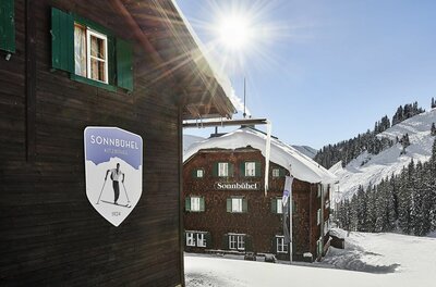 Holzhütte Sonnbühel im Schnee mit Logo an der Fassade und verschneiten Bergen im Hintergrund bei Sonnenschein.