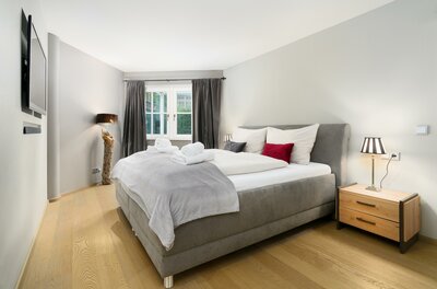 Schlafzimmer Kitz-Heart Premium Modernes Schlafzimmer mit grauem Polsterbett, Holzparkett, Nachttisch und Fenster mit dunklen Vorhängen.