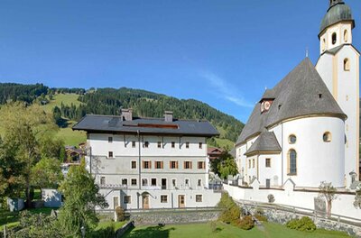 Mehrstöckiges weißes Gebäude neben einer Kirche mit Zwiebelturm vor bewaldeten Bergen.
