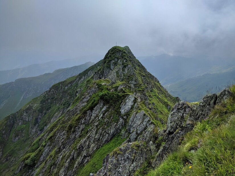 Steiler, felsiger Berggipfel mit grünem Bewuchs unter bewölktem Himmel.