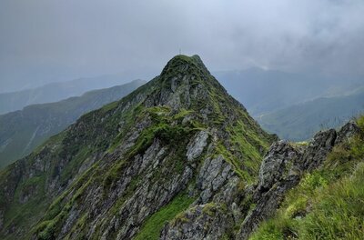 Steiler, felsiger Berggipfel mit grünem Bewuchs unter bewölktem Himmel.