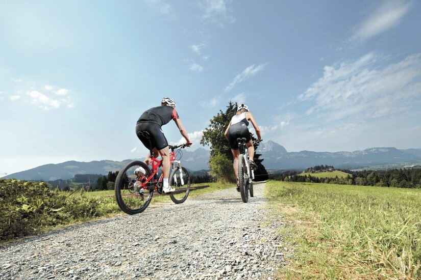 Mountainbiken in Kitzbühel Zwei Mountainbiker fahren von hinten fotografiert auf einem Schotterweg durch eine Wiesenlandschaft vor Bergkulisse.