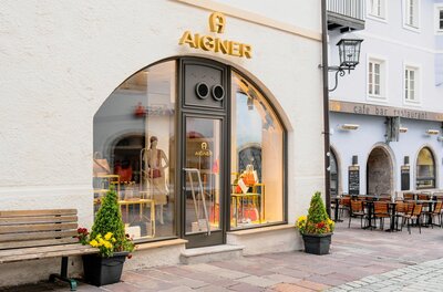 Aigner Shop Außenansicht Außenansicht des Aigner Shops mit Schaufenster, Taschen und einer Parkbank davor.