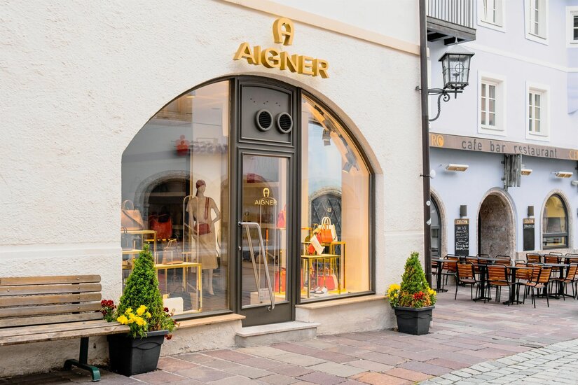 Außenansicht des Aigner Shops mit Schaufenster, Taschen und einer Parkbank davor.