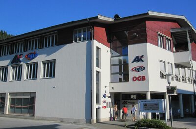 BFI Kitzbühel Ein modernes Gebäude mit dem Logo der AK und OGB. Es ist klarer Himmel und sonniges Wetter zu sehen.