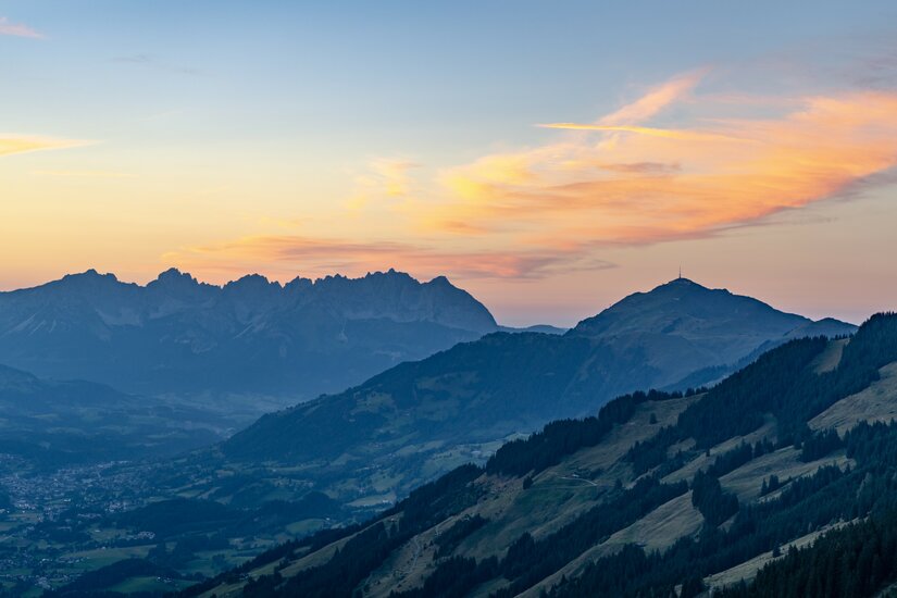 Blick über bewaldete Berghänge auf ein gezacktes Bergmassiv unter einem Abendhimmel mit orangefarbenen Wolken.