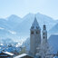 Die idyllische Winterlandschaft zeigt eine charmante Stadt mit Kirchen und schneebedeckten Bäumen, umgeben von majestätischen Alpen.