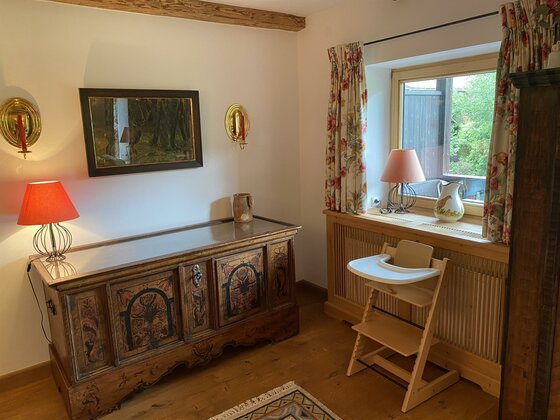Innenansicht Ferienwohnung Schmachtl Rustikaler Innenraum mit bemalter Holztruhe, Kinderhochstuhl und Fenster mit Blumenmustervorhängen.