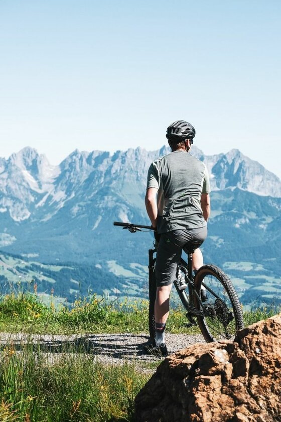Mountainbiker vor Bergpanorama Ein Mountainbiker blickt von einem Schotterweg auf ein markantes Gebirgsmassiv unter blauem Himmel.