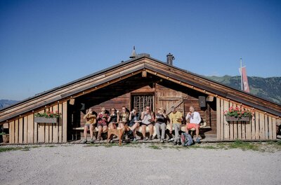 Rast an der Almhütte Eine Gruppe von Wanderern sitzt mit einem Hund auf einer Holzbank vor einer rustikalen Almhütte unter blauem Himmel.