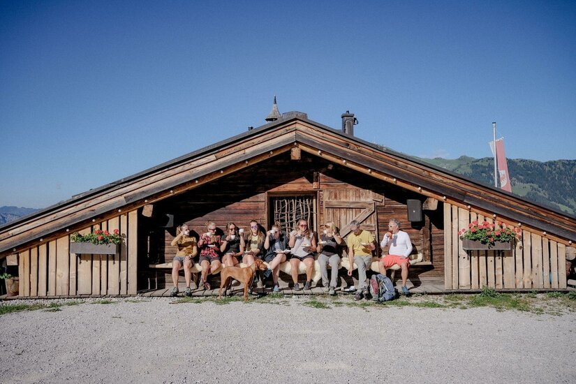 Rast an der Almhütte Eine Gruppe von Wanderern sitzt mit einem Hund auf einer Holzbank vor einer rustikalen Almhütte unter blauem Himmel.
