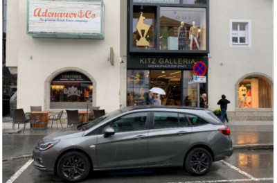 Ein graues Auto parkt vor Geschäften in einer Stadt. Der Himmel ist bewölkt und es regnet leicht.