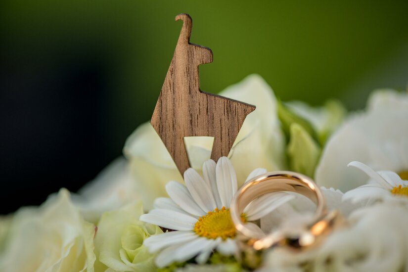 Hochzeit <p>Eine Holzfigur in Form einer Gams steht auf einer bunten Blumenarrangierung. Im Vordergrund sind goldene Ringe zusammen mit weißen Margeriten zu sehen.</p>