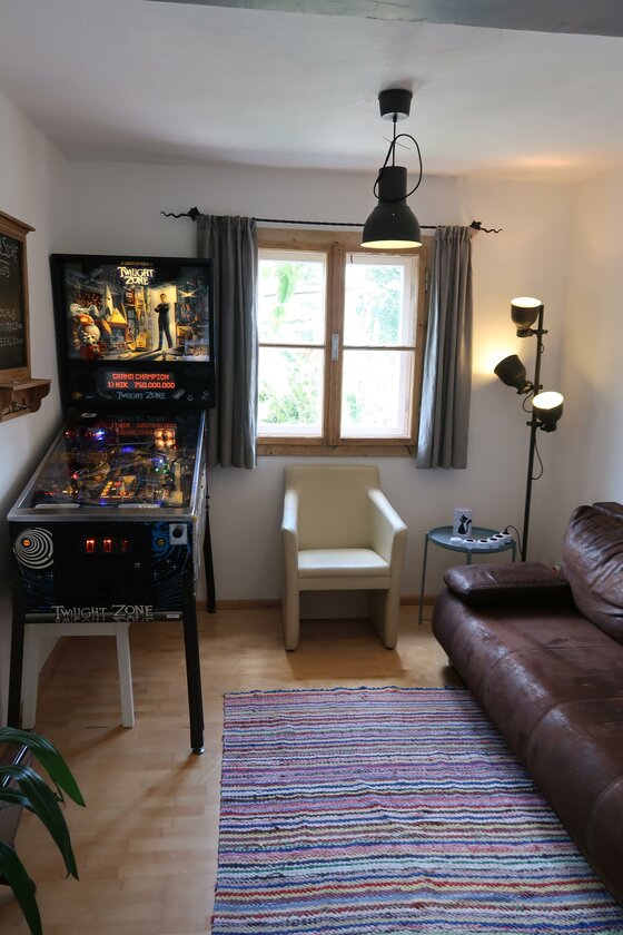 Retro Apartment Kitzbühel Innenansicht eines Zimmers mit Flipperautomat, Ledersofa und hellem Sessel vor einem Fenster.