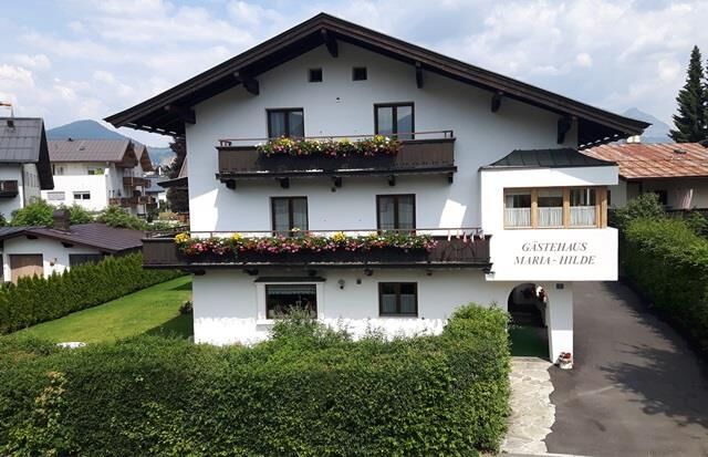 Sommeransicht Gästehaus Maria Hilde Vorderansicht eines weißen Gästehauses mit dunklen Holzbalkonen und Blumenschmuck.