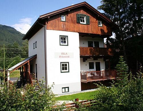 Villa Schönfeld im Sommer Dreistöckiges weißes Gebäude mit Holzgiebel und Balkonen vor bewaldetem Berg.