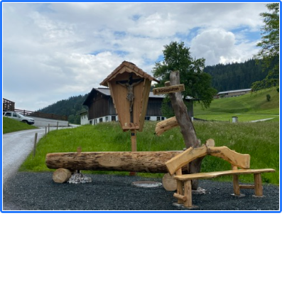 Rastplatz mit Holzbrunnen Ein Rastplatz mit Holzbank, Brunnen aus einem Baumstamm und einem Kruzifix vor einer Bergkulisse.
