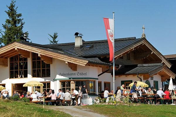 Hahnenkammstüberl im Sommer Außenansicht des Hahnenkamm Stüberls mit Gästen auf der Terrasse und Tiroler Fahne unter blauem Himmel.