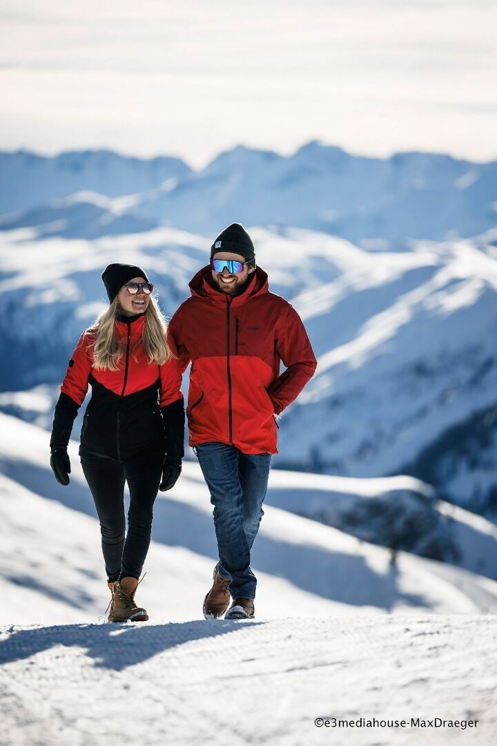 Winterwandern in den Kitzbüheler Alpen Ein Paar in Winterkleidung wandert auf einem verschneiten Bergpfad vor einem alpinen Panorama.