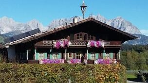 Hölzlhof-Vermietung-Bauernhof-Kitzbühel-Sommer-Hau Ein traditionelles alpenländisches Haus mit bunten Blumen auf den Balkonen. Im Hintergrund sind majestätische Berge und ein klarer blauer Himmel zu sehen.