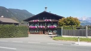 Hölzlhof-Vermietung-Bauernhof-Kitzbühel-Sommer Ein traditionelles Alpenhaus mit bunten Blumen an den Fenstern. Im Hintergrund sind Berge zu sehen.