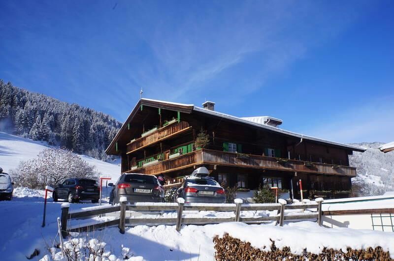 MyApart 1709 Jochberg im Winter Großes Holzhaus im alpinen Stil mit Balkonen und parkenden Autos in einer verschneiten Winterlandschaft.