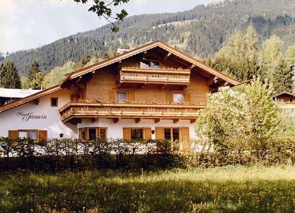 Haus Jasmin im Sommer Zweistöckiges Alpenhaus aus Holz und weißem Putz mit Blumenbalkonen vor bewaldetem Berg.
